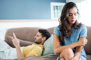 man laying on sofa on phone and woman sitting on edge of sofa looking disappointed