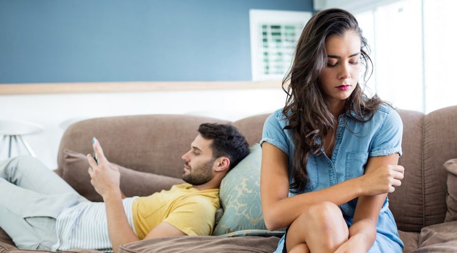 man laying on sofa on phone and woman sitting on edge of sofa looking disappointed