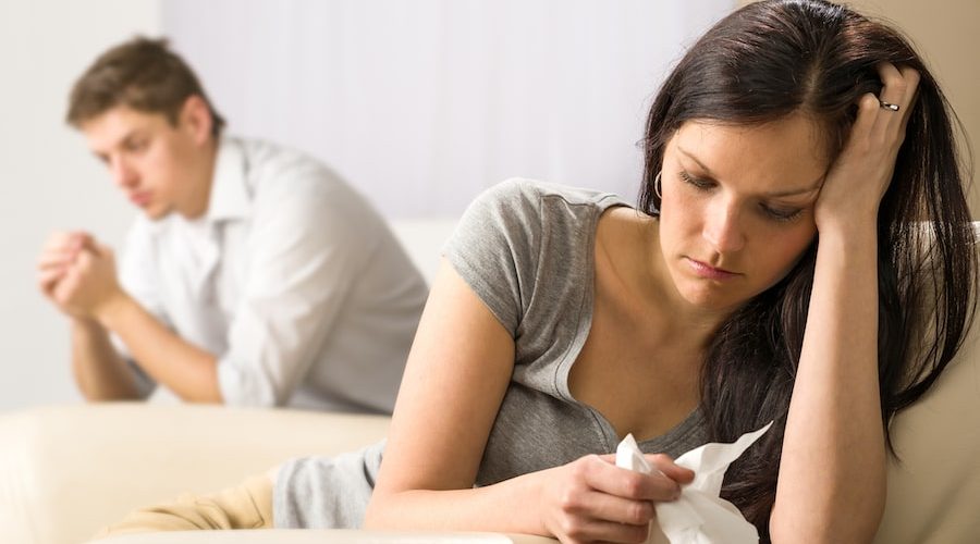 couple looking away from each other on the sofa, woman holding tissue and man looking serious