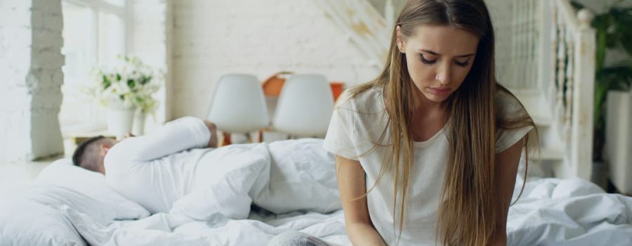 woman-sitting-on-bed-man-sleeping-behind-her