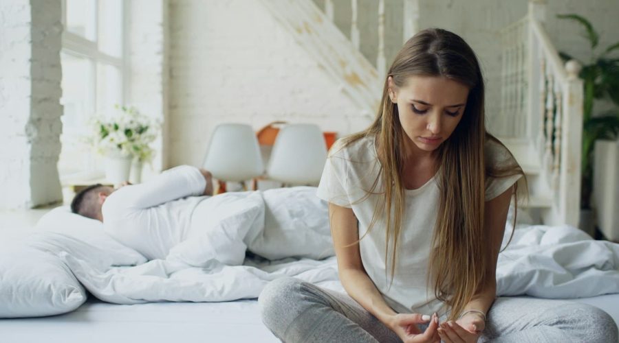 woman-sitting-on-bed-man-sleeping-behind-her